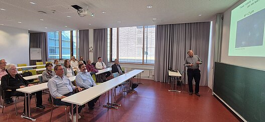 Prof. Dr. Peer Fischer erläutert einer sitzenden Besuchergruppe Forschungsinhalte anhand einer Präsentation.