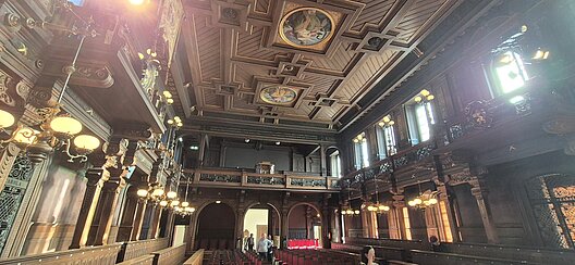 Innenansicht der historischen Alten Aula der Universität Heidelberg mit reich verzierter Holzdecke, Emporen und Wandmalereien.