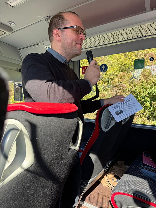 Der VHV-Vorsitzende spricht während der Busfahrt nach Heidelberg mit einem Mikrofon zu den Teilnehmerinnen und Teilnehmern.